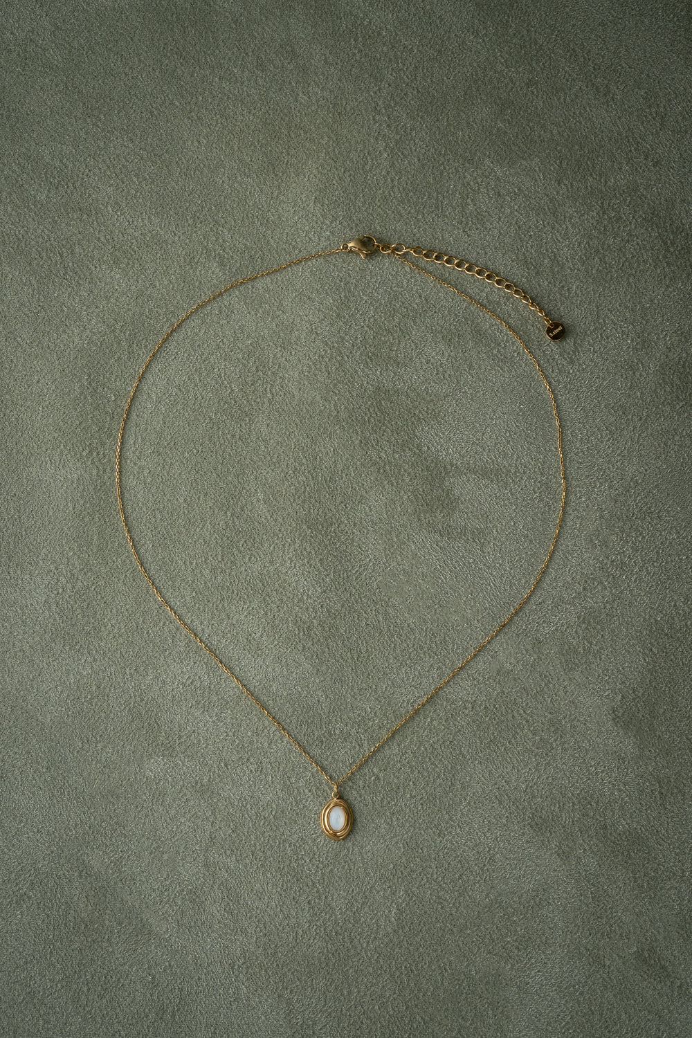 A delicate gold chain necklace with a small teardrop-shaped mother of pearl pendant featuring a white inlay, laid flat on a textured gray background.