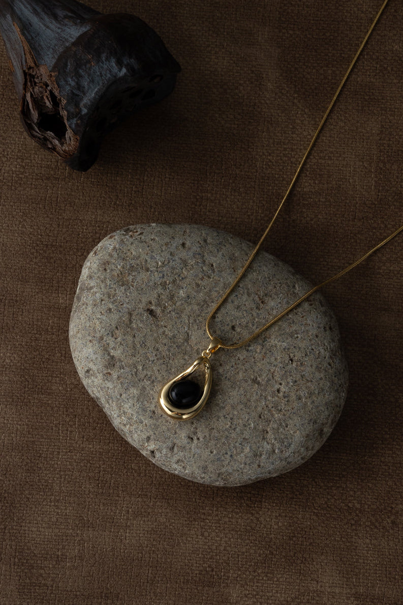 A gold teardrop pendant necklace with a dark stone center, draped over a smooth gray stone on a textured brown fabric background.