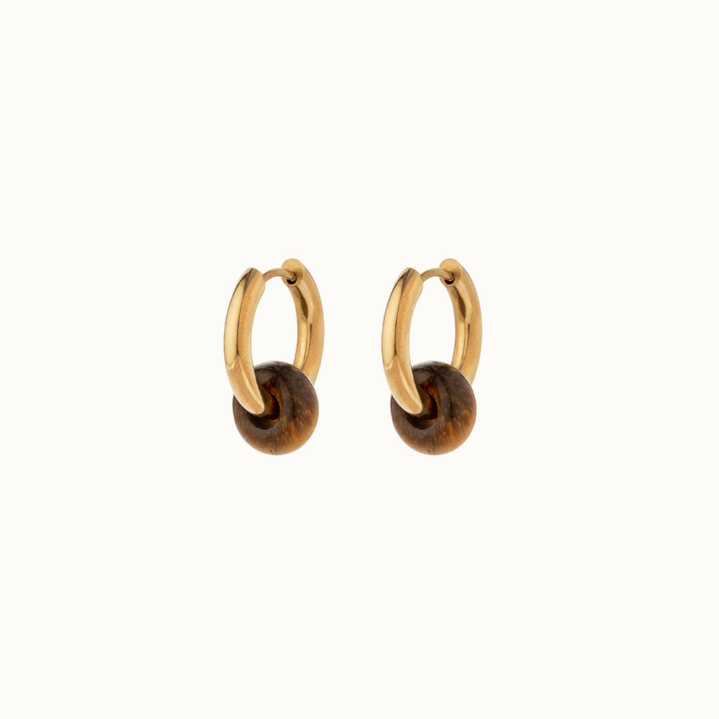 A pair of gold huggie hoop earrings with a polished brown tiger's eye bead dangling from the bottom, shown on a clean off-white background.