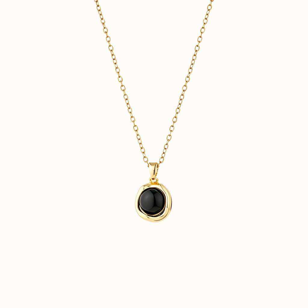 A delicate gold chain necklace with a small, irregularly shaped black onyx stone pendant in a gold bezel setting, shown against a plain off-white background.