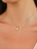 Close-up of a woman wearing a delicate gold chain necklace with a small round gold Mother of Pearl pendant, paired with a matching gold stud earring, against a warm beige background.