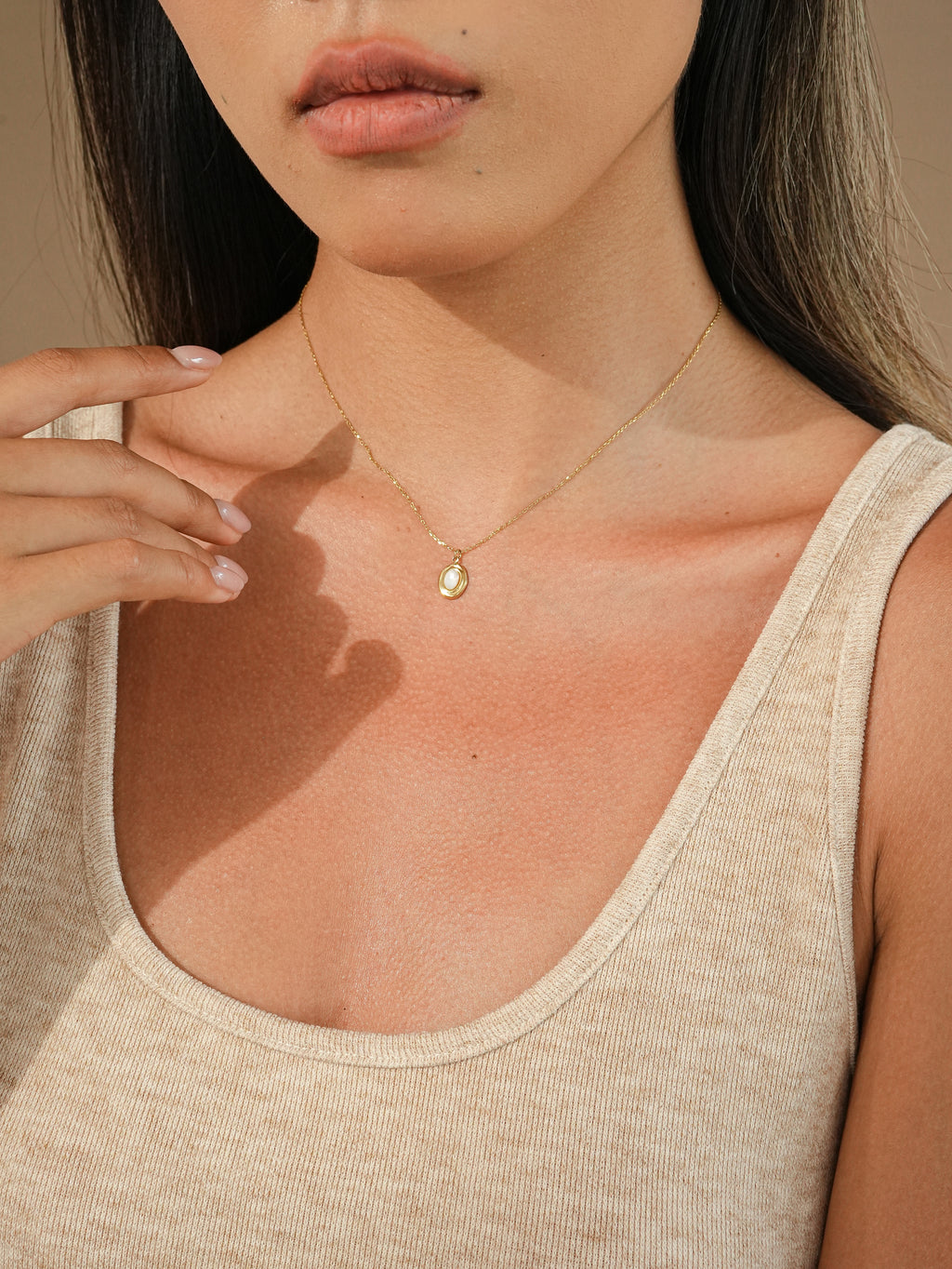 A close-up of a woman wearing a delicate gold chain necklace with a small teardrop-shaped gold mother of pearl pendant, styled with a beige ribbed tank top.