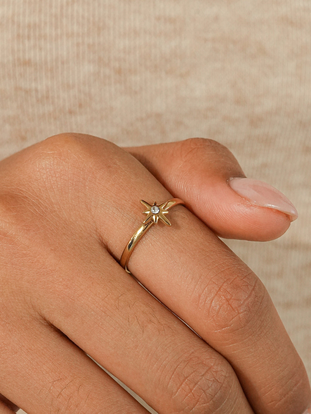 A close-up of a hand wearing a delicate gold ring with a small starburst cluster of clear stones, set against a soft beige knit background.