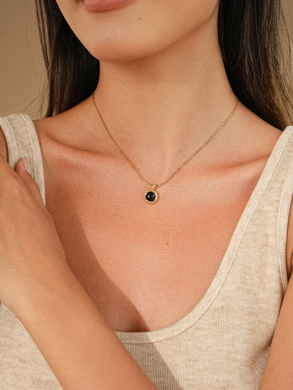 A close-up of a model wearing a delicate gold chain necklace with a small round pendant featuring a dark green stone, paired with a light beige ribbed tank top.