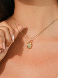 A close-up of a woman wearing a delicate gold chain necklace with an oval pendant featuring a pale grey cat's eye stone, paired with a beige ribbed tank top.
