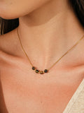 A close-up of a model wearing a delicate gold necklace with a small cluster of dark brown and black beads at the center, styled with a beige ribbed tank top.