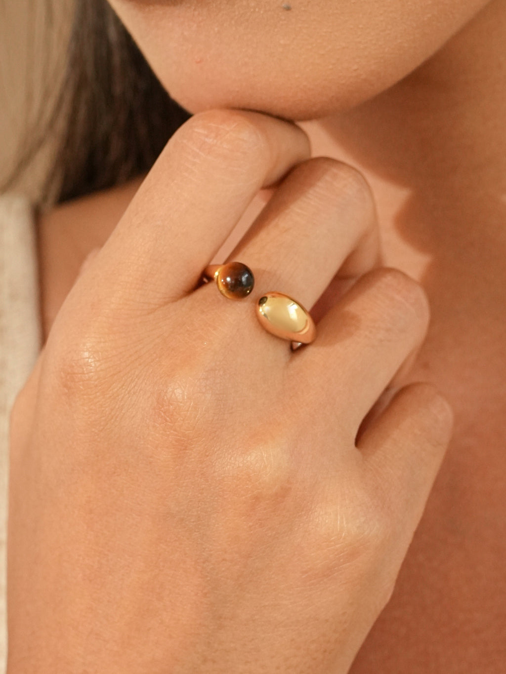 A close-up of a model's hand wearing adjustable gold rings, one with a small dark brown tiger eye natural stone crystal bead and the other with a polished oval gold setting, against a warm beige background.