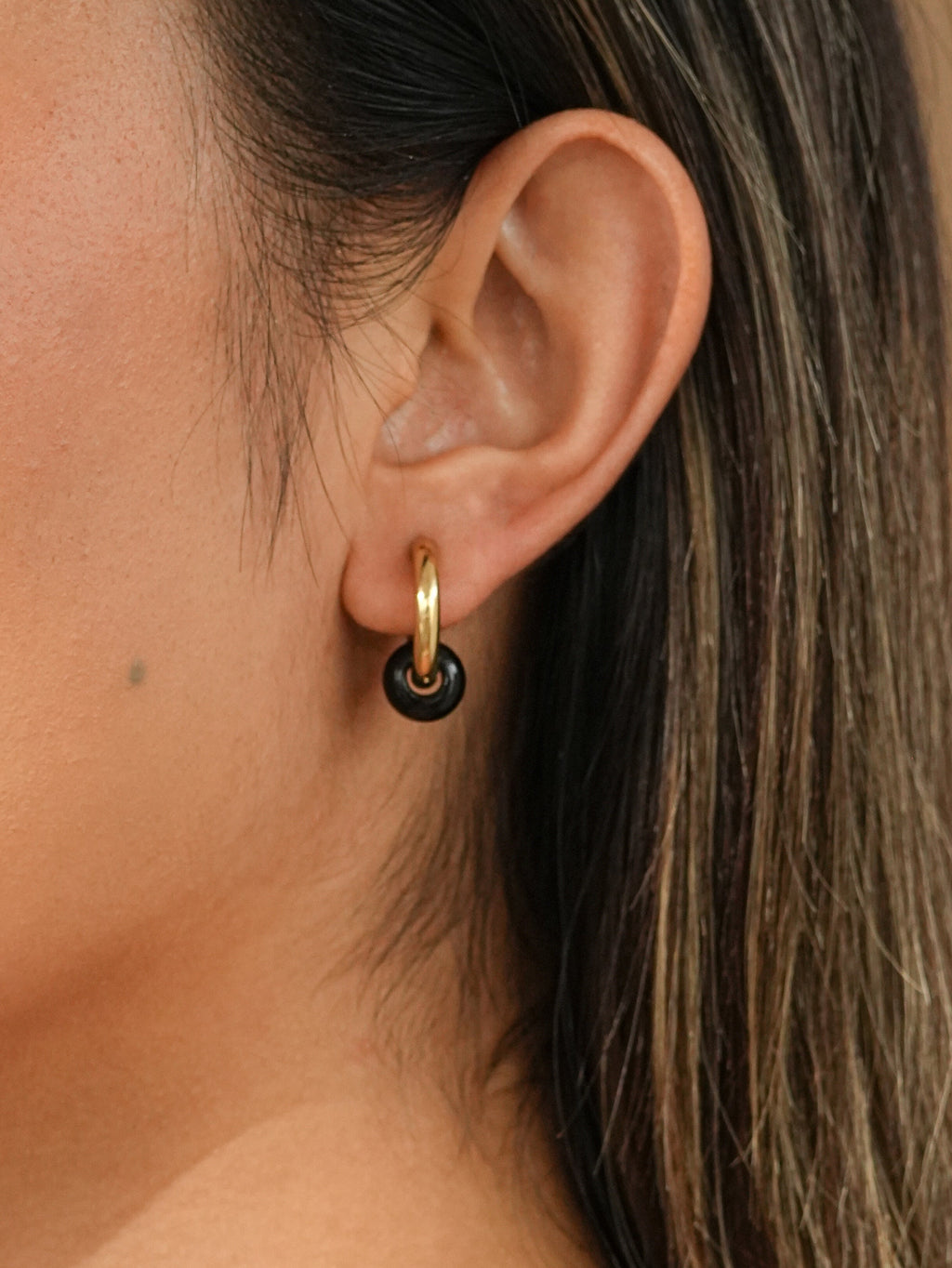 A close-up side profile of a woman wearing a small gold hoop earring with a dark brown tiger eye natural crystal circular charm, against a warm beige background.