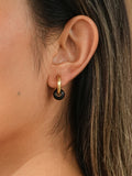 A close-up side profile of a woman wearing a small gold hoop earring with a dark brown tiger eye natural crystal circular charm, against a warm beige background.